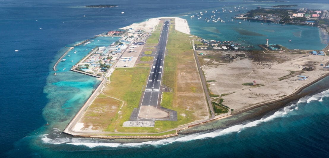 Luftaufnahme eines schmalen Insel-Flughafens mit einer Landebahn, umgeben von Meer und Booten