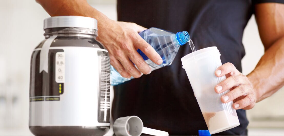 Mann gießt Wasser aus einer Flasche in einen Protein-Shaker neben einem großen Behälter mit Proteinpulver