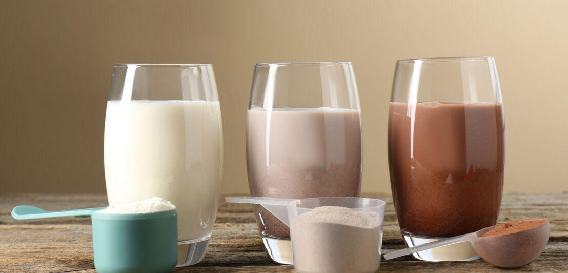 Drei Gläser mit weißem, hellbraunem und dunklem Proteinshake, davor drei Löffel mit weißem, hellbraunem und dunklem Proteinpulver auf Holztisch
