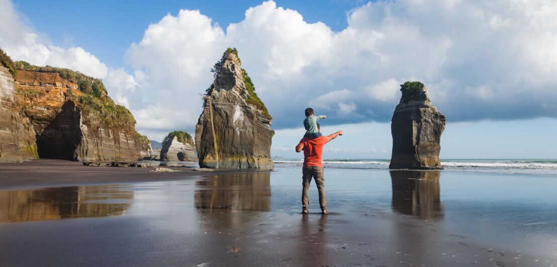 Vater trägt seinen kleinen Sohn auf der Schulter, beide bestaunen die felsige Küstenlandschaft des Three Sister Beach in North Taranaki.