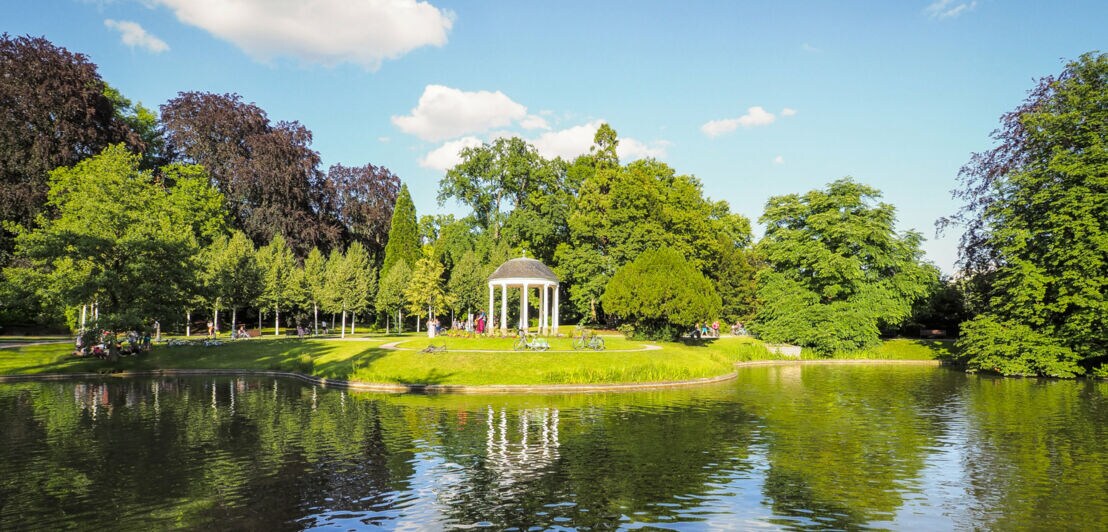 Parkanlage mit einem weißen Pavillon am Ufer eines Sees unter blauem Himmel mit Wolken