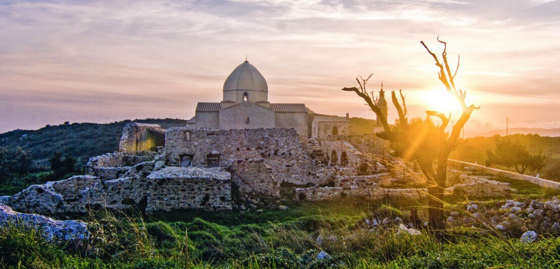 Sonnenuntergang über einer alten Steinruine mit einer Kuppel und einem kahlen Baum im Vordergrund