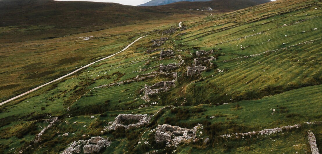 Landschaft mit grünen Wiesen und Überresten von Steinhäusern, die sich entlang eines Pfades erstrecken. Im Hintergrund sind sanfte Hügel sichtbar.