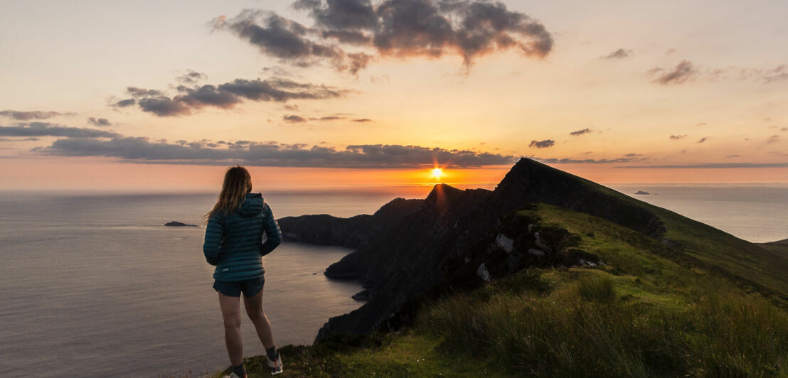 Eine Frau mit langen Haaren steht auf einem Hügel und schaut auf einen Sonnenuntergang über dem Meer. Sie trägt eine grüne Jacke und Shorts. Im Hintergrund sind dunkle Felsen und der Horizont sichtbar.