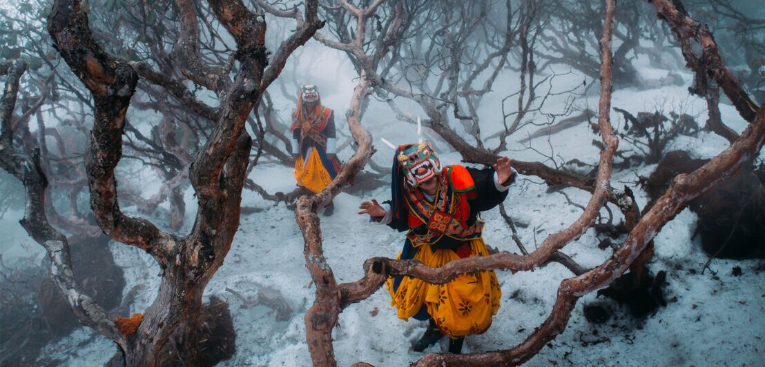 Zwei Personen mit asiatischen Drachenmasken stehen inmitten schneebedeckter Bäume.