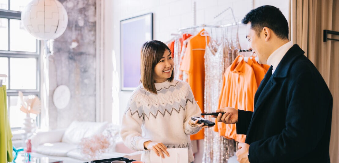 Fotografie eines Mannes, der in einer Boutique seinen Einkauf via Smartphone an einem mobilen Kartengerät bezahlt.