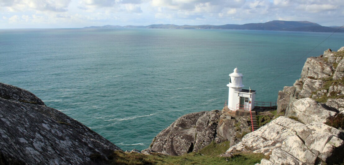 Ein weißer Leuchtturm steht auf einer Klippe über dem Meer, umgeben von Felsen und Gras. Der Himmel ist bewölkt mit einigen blauen Abschnitten.