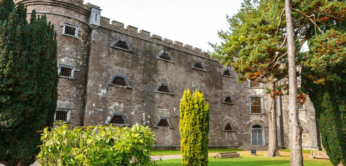 Historisches Gebäude mit steiniger Fassade und Fenster mit Gittern, umgeben von Bäumen und Rasenflächen.