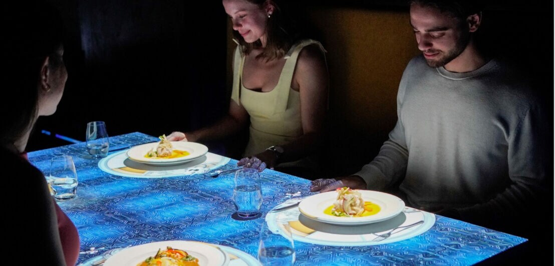 Drei Personen sitzen an einem Tisch mit Projektion. Auf dem Tisch sind zwei Teller mit einer Speise und Gläser sichtbar. Der Tisch hat ein blaues, gemustertes Design