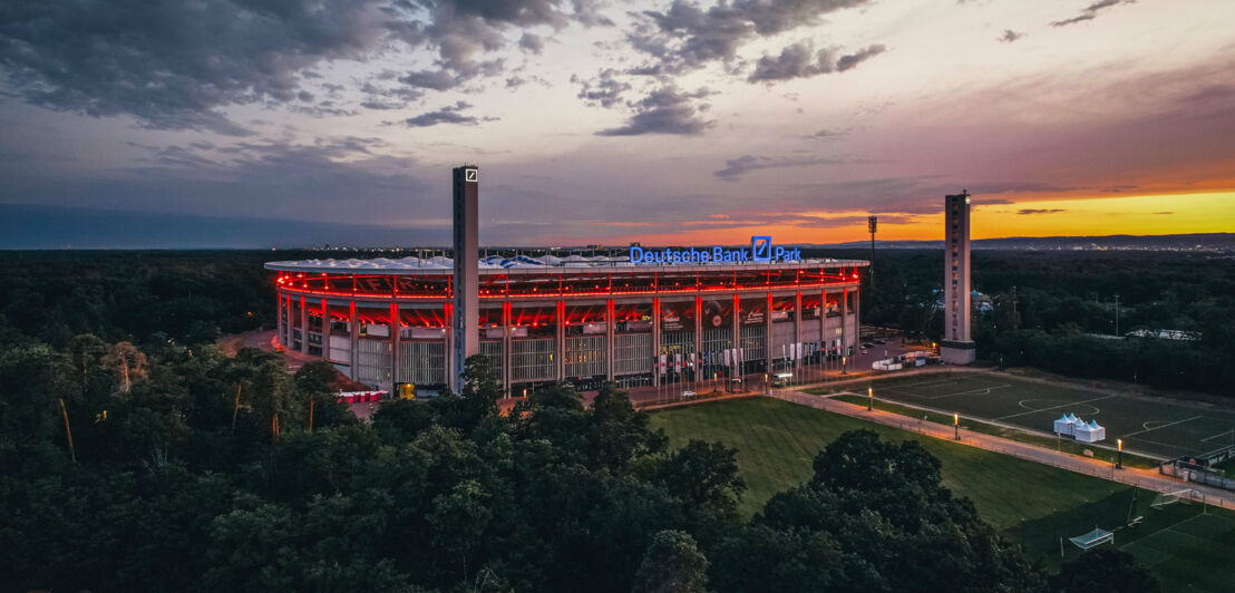 Rot beleuchtetes Stadion mit Deutsche-Bank-Logo in einem Waldgebiet bei Dunkelheit.