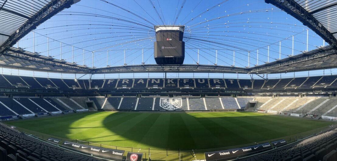 Innenansicht eines Fußballstadions mit Schriftzug Frankfurt und Adler-Wappen auf menschenleeren Rängen unter blauem Himmel.