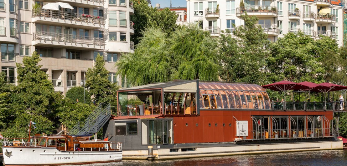 Ein Restaurantboot mit großen Fenstern und einem roten Dach liegt am Ufer eines Flusses. Im Hintergrund sind moderne Wohngebäude und Bäume sichtbar.