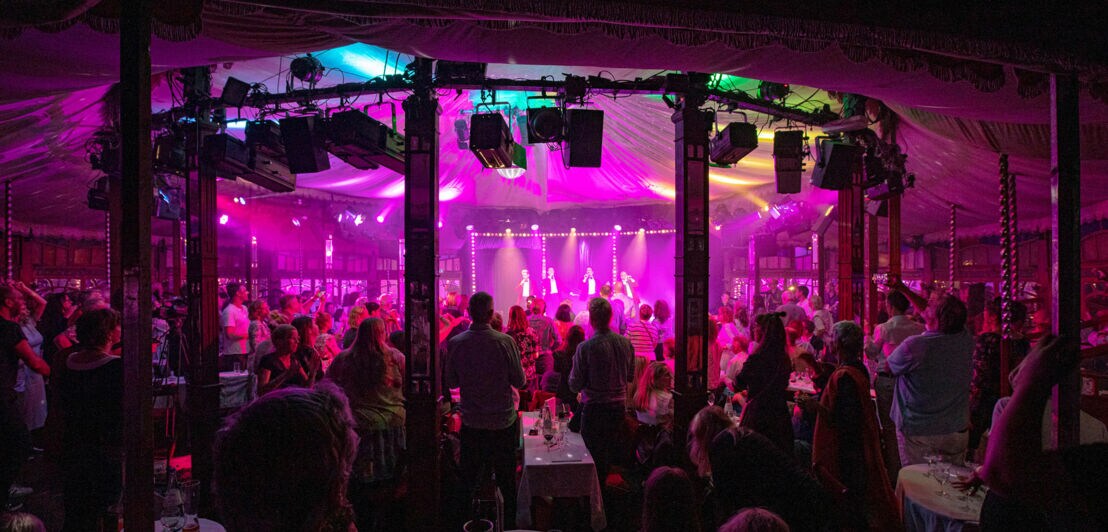 Eine Menschenmenge steht in einem Zelt mit bunten Lichtern. Auf der Bühne sind mehrere Personen sichtbar, die performen. Tische sind im Vordergrund.