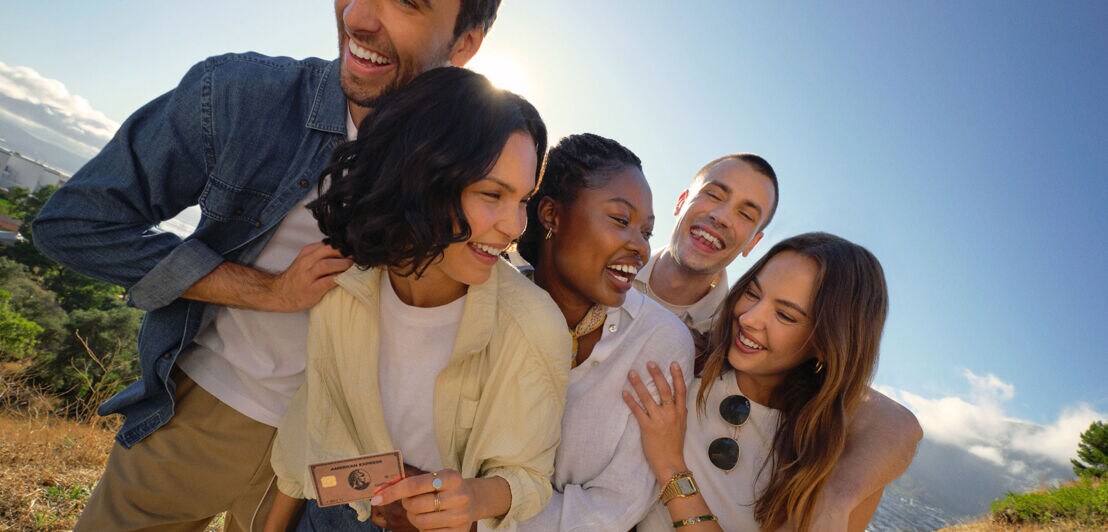 Drei Frauen und zwei Männer liegen sich lachend in den Armen und stehen bei strahlendem Sonnenschein auf einem Weg in der Natur.