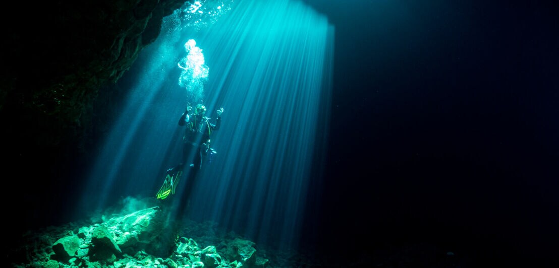 Ein Taucher schwebt in einem dunklen Unterwasserraum, umgeben von Lichtstrahlen, die von der Oberfläche kommen. Der Boden besteht aus Steinen.