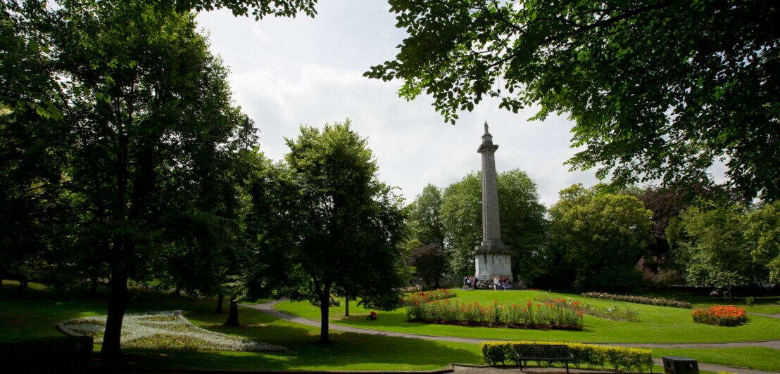 Blick auf den People's Park in Limerick City mit einem hohen Obelisken in der Mitte, umgeben von Bäumen und Blumenbeeten.
