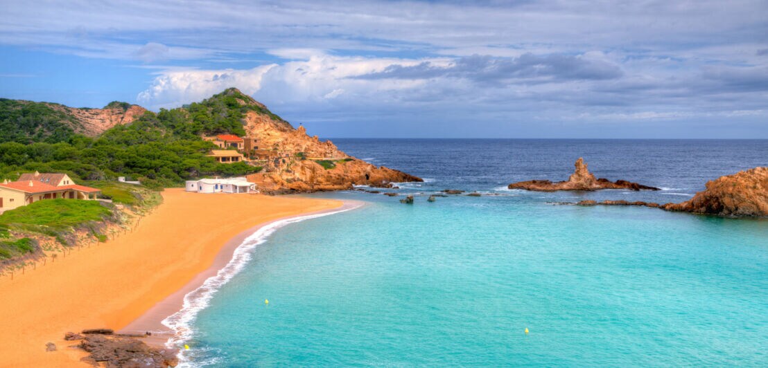 Strand mit feinem Sand und klarem Wasser, umgeben von grünen Hügeln und Felsen. Im Hintergrund sind einige Häuser sichtbar.