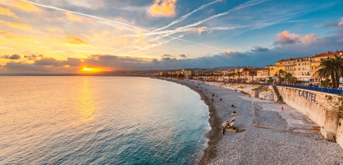 Blick auf die Küste von Nizza bei Sonnenuntergang, mit einem Kiesstrand, Palmen und Gebäuden entlang der Uferpromenade.