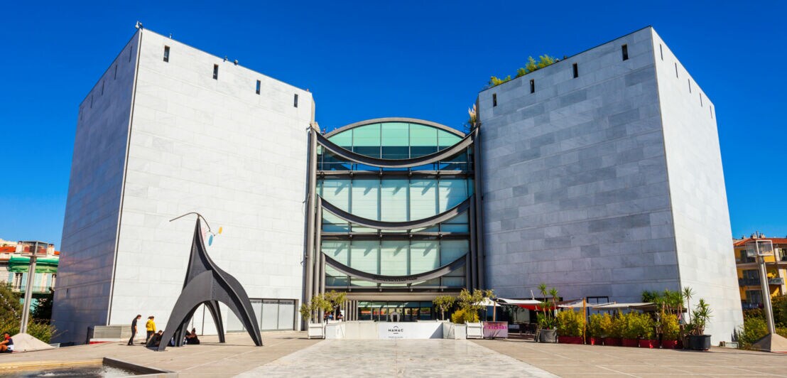 Moderne Architektur eines Gebäudes mit zwei großen, rechteckigen Türmen aus hellem Stein. Vor dem Gebäude steht eine schwarze Skulptur. Klare blaue Himmel.