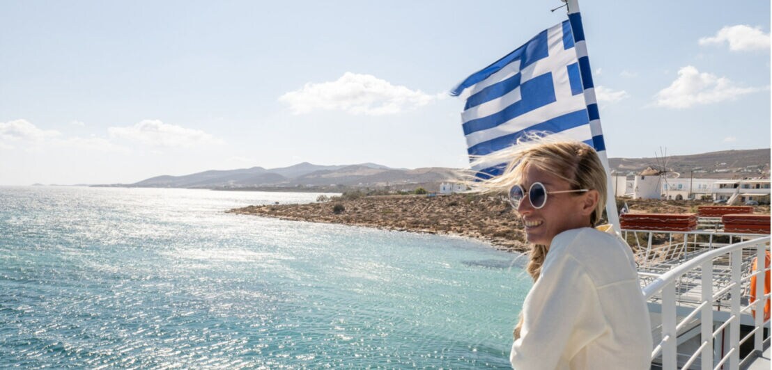 Person lehnt am Geländer einer Fähre mit wehender griechischer Flagge, Blick auf das blaue Meer und eine felsige Küste im Hintergrund