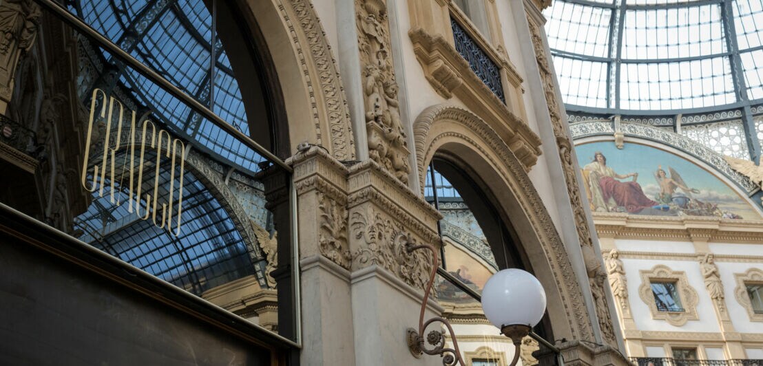 Architekturansicht eines eleganten Gebäudes mit großen Bögen und kunstvollen Verzierungen. Reflektierende Fenster zeigen den Innenraum und eine Wandmalerei.