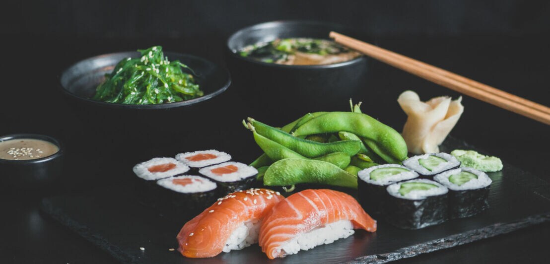 Ein schwarzes Servierbrett mit Sushi, darunter zwei Lachs-Sashimi-Stücke, eine Rolle mit Lachs und Avocado, Edamame, Algensalat, eingelegtem Ingwer und Miso-Suppe in Schalen. Essstäbchen liegen daneben.