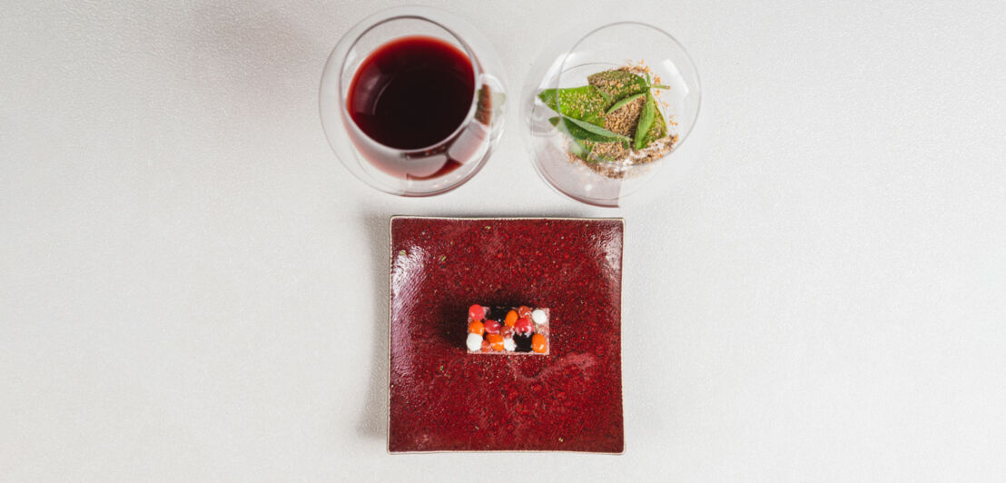 Ein quadratischer, roter Teller mit einem kleinen Dessert, das aus mehreren bunten Schichten besteht. Daneben stehen zwei Gläser: eines mit rotem Getränk und eines mit einer grünen Speise.