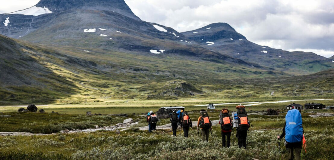 Gruppe von sieben Wanderern mit Rucksäcken auf einem Wanderweg in einer bergigen Landschaft. Im Hintergrund sind hohe Berge und bewölkter Himmel sichtbar.