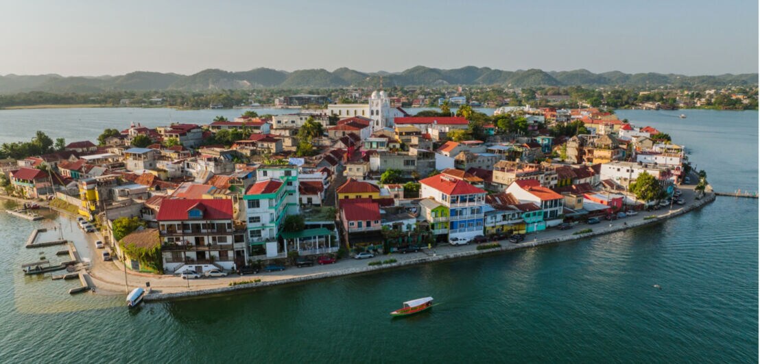 Luftaufnahme einer kleinen Insel mit dicht bebauten Häusern mit roten Dächern, umgeben von Wasser und Bergen im Hintergrund