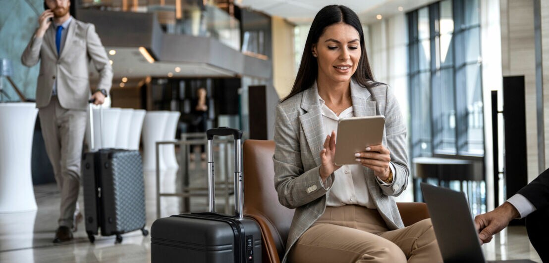 Eine Frau in heller Kleidung sitzt in einer Flughafen-Lounge und nutzt ein Tablet, im Hintergrund geht ein Mann mit Telefon am Ohr im grauen Anzug mit Trolley.