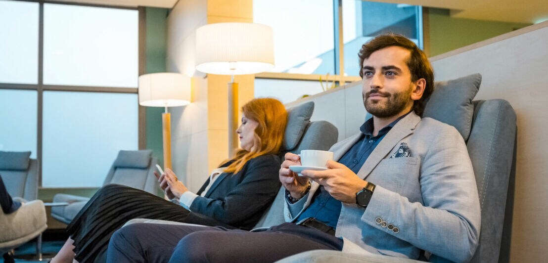 Zwei elegant gekleidete Personen sitzen entspannt in Sesseln einer Flughafen-Lounge mit Panoramafenster, eine hält eine Tasse Kaffee, die andere schaut auf ihr Smartphone.