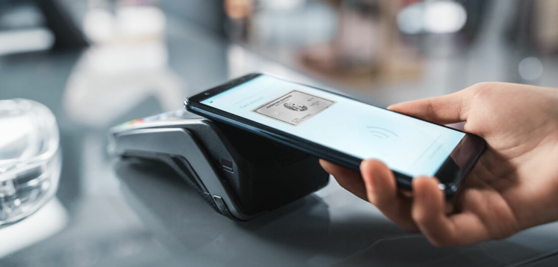 Eine Person hält ein Smartphone, dessen Display die Business Platinum Card von American Express zeigt, an ein Kartenlesegerät.