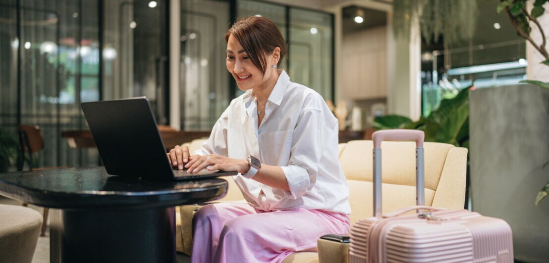 Eine Frau sitzt lächelnd an einem Laptop in einer Lounge, neben ihr ein großer rosafarbener Hartschalenkoffer.