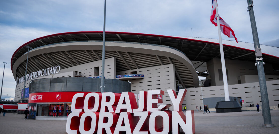 Das Estadio Metropolitano mit dem Schriftzug 'CORAJÉ Y CORAZÓN' im Vordergrund. Im Hintergrund sind Flaggen und die Stadionarchitektur sichtbar.