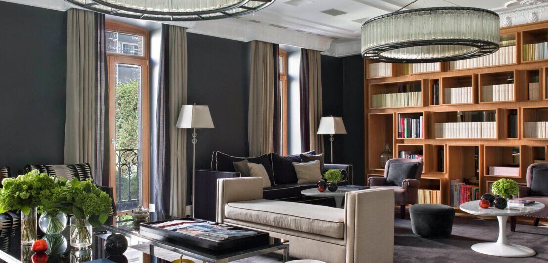 Hotelbibliothek im Hotel Unico Madrid mit zwei großen, runden Kronleuchtern, einem beigen Sofa, einem Glastisch und einer Bücherwand. Pflanzen und Dekorationen auf dem Tisch.