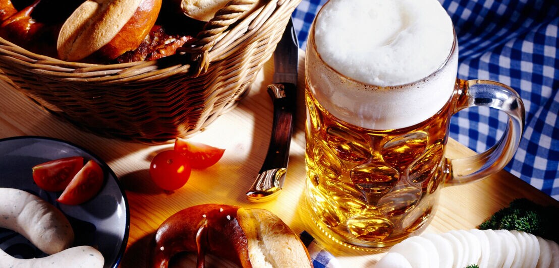Ein Korb mit Brötchen, ein Glas Bier mit Schaum, eine Brezel, Wurst, Tomaten, Senf und Käse auf einem Holztisch mit kariertem Tuch.