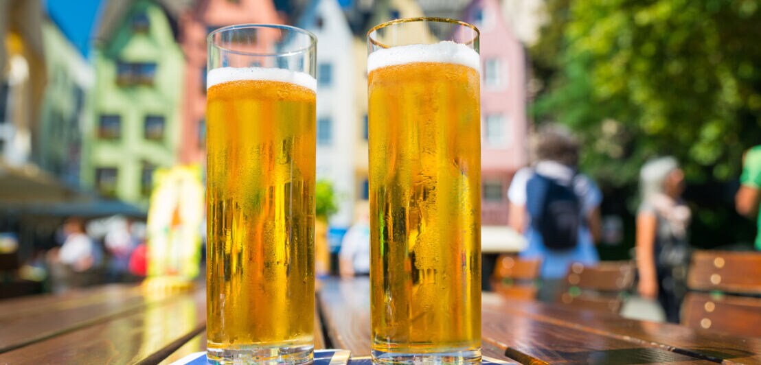Zwei Gläser Bier auf einem Holztisch, im Hintergrund farbenfrohe Gebäude und Bäume. Die Gläser sind transparent mit goldenem Bier und Schaumkrone.