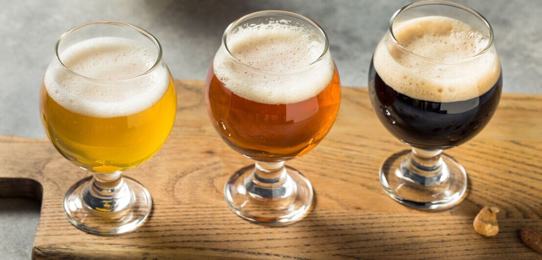 Drei Gläser mit verschiedenen Biersorten auf einem Holzbrett. Links ein helles Bier, in der Mitte ein bernsteinfarbenes Bier und rechts ein dunkles Bier. Schaumkrone auf jedem Glas.