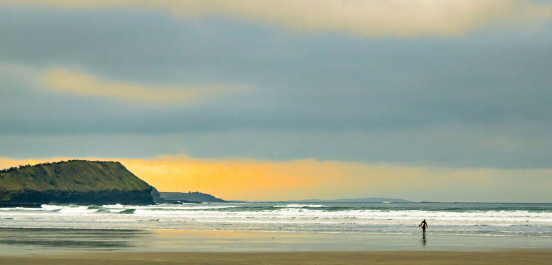 Eine Person steht im flachen Wasser am Strand, während die Wellen sanft an den Ufer schlagen. Im Hintergrund ist eine bewaldete Küstenlinie und ein bewölkter Himmel zu sehen.