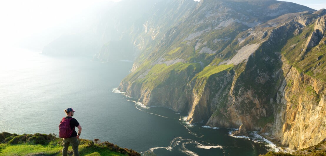 Ein Wanderer mit einem Rucksack steht auf einer grünen Wiese und blickt auf eine steile Küste mit Klippen und dem Meer im Hintergrund.