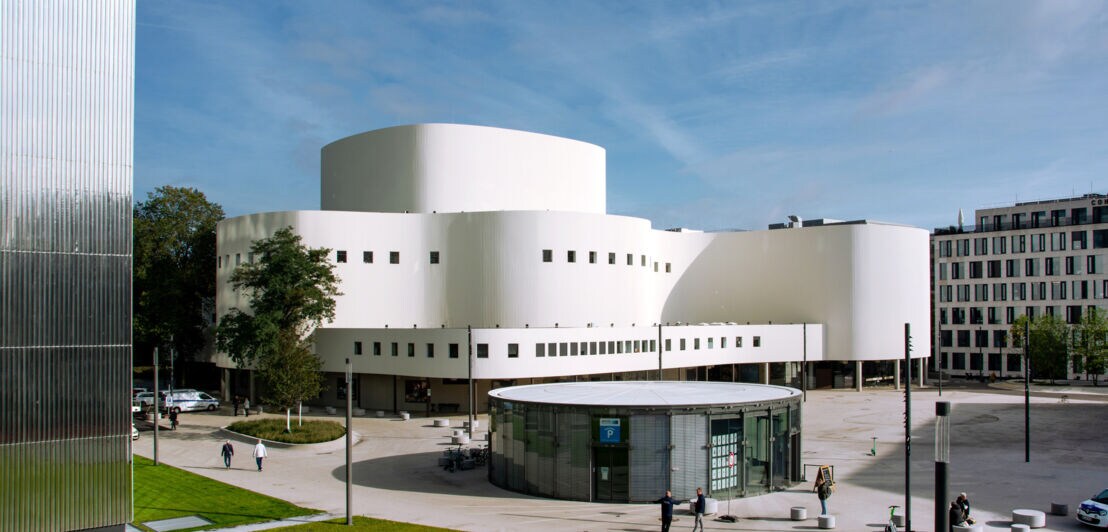Modernes weißes Theatergebäude mit runden Formen und kleinen quadratischen Fenstern, umgeben von einem Platz mit wenigen Menschen und Autos