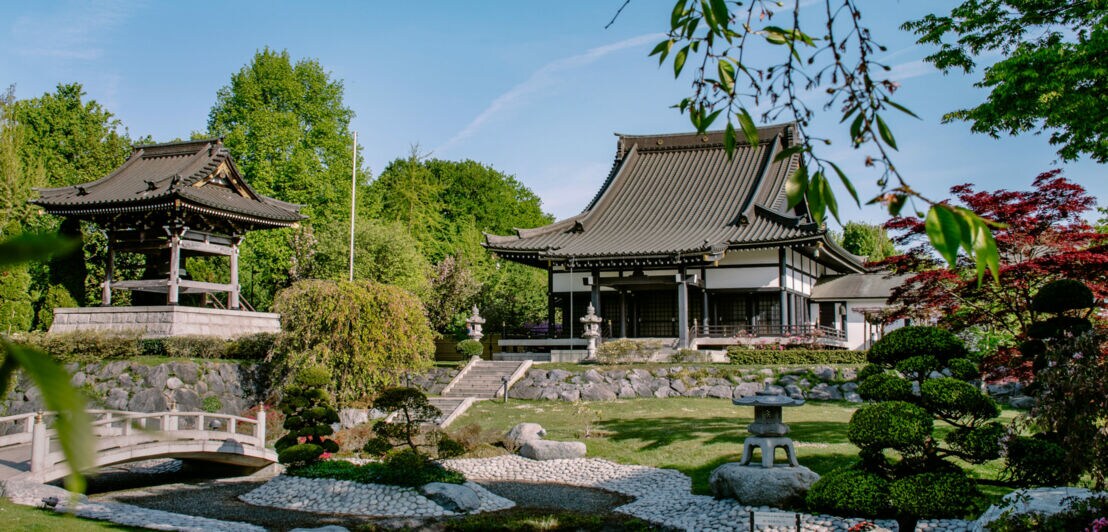 Japanischer Garten mit zwei traditionellen Gebäuden, einem Pavillon und einem Haus mit geschwungenem Dach. Grüne Bäume und gepflegte Sträucher umgeben den Garten.