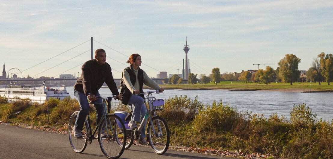 Zwei Personen auf Fahrrädern fahren am Ufer eines Flusses entlang. Im Hintergrund sind Bäume, ein Fernsehturm und ein Riesenrad sichtbar.