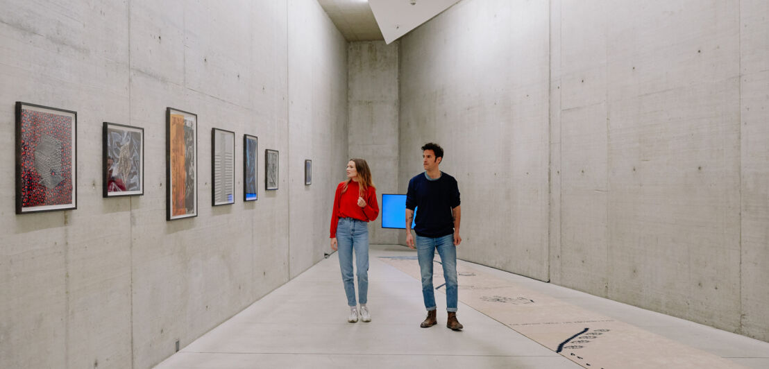 Zwei Personen in einem Ausstellungsraum der Kunst im Tunnel Düsseldorf. Links hängen mehrere Bilder an der Wand, der Boden zeigt eine grafische Darstellung.