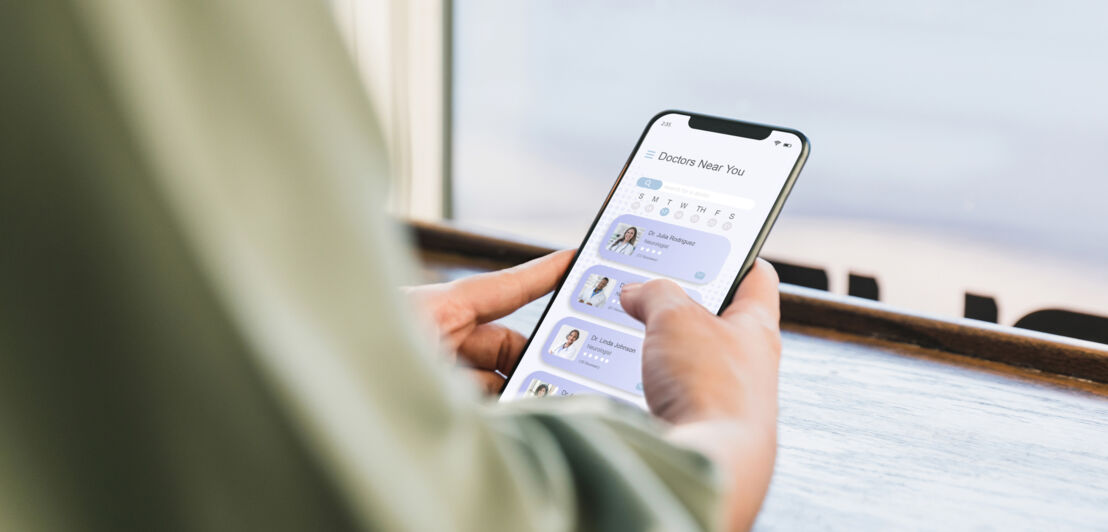 Eine Person hält ein Smartphone in der Hand und scrollt durch eine App mit Profilbildern und Namen. Auf dem Tisch steht ein Glas mit kaltem Kaffee.