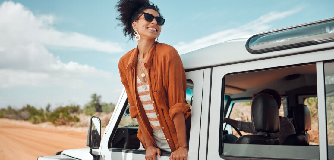 Eine Frau mit lockigem Haar lehnt aus dem Fenster eines weißen Geländewagens. Sie trägt eine orangefarbene Bluse und eine gestreifte Bluse darunter. Im Hintergrund ist eine Landschaft mit Bäumen und einem blauen Himmel zu sehen.