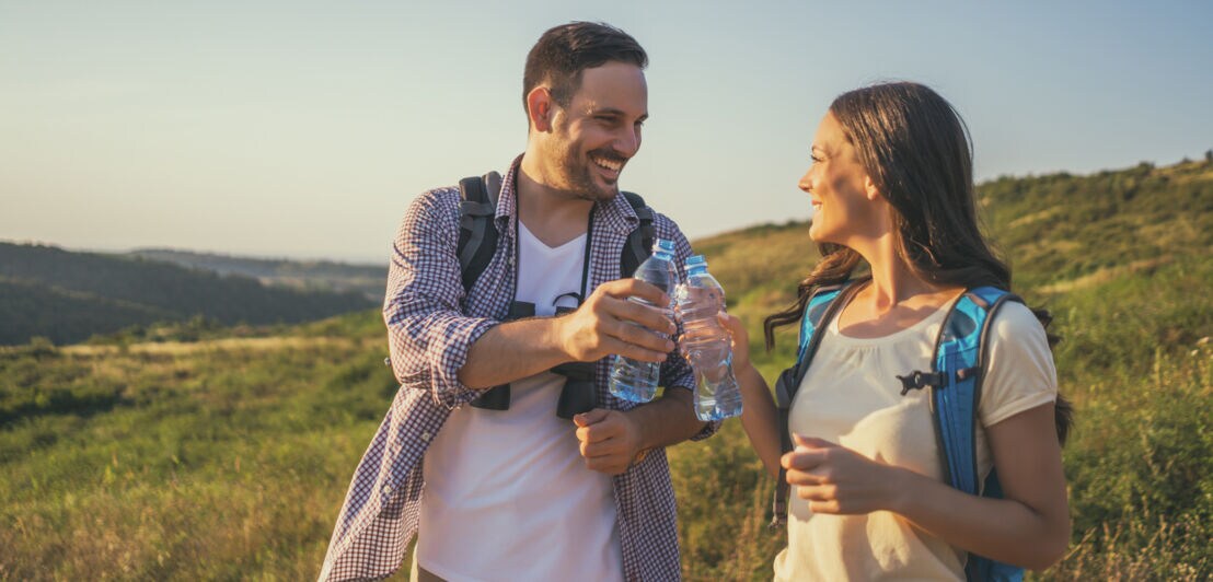 Zwei Personen stehen auf einer Wiese, der Mann reicht der Frau eine Wasserflasche. Beide tragen Rucksäcke und lächeln.