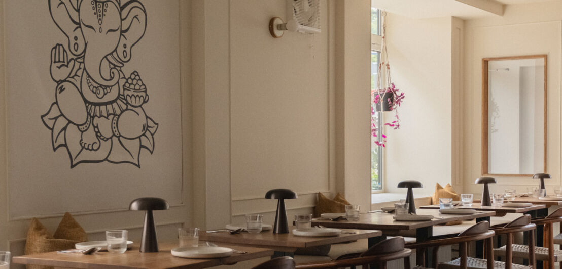 in Restaurant mit Holztischen und Stühlen. An der Wand ist eine große, schwarze Darstellung von Ganesha. Es gibt Fenster mit natürlichem Licht.