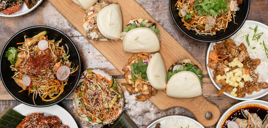 Eine Vielzahl von Gerichten auf einem Tisch: zwei Schalen mit gebratenen Nudeln, drei Schalen mit verschiedenen Fleischgerichten, Bao-Brötchen, Frühlingsrollen und Desserts.