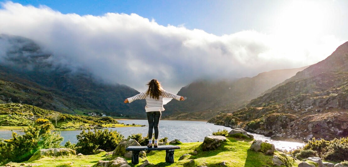 Eine Person mit langen, offenen Haaren steht auf einem Stein und breitet die Arme aus. Sie trägt ein gestreiftes Oberteil und blickt auf einen See, umgeben von Bergen und Wolken.